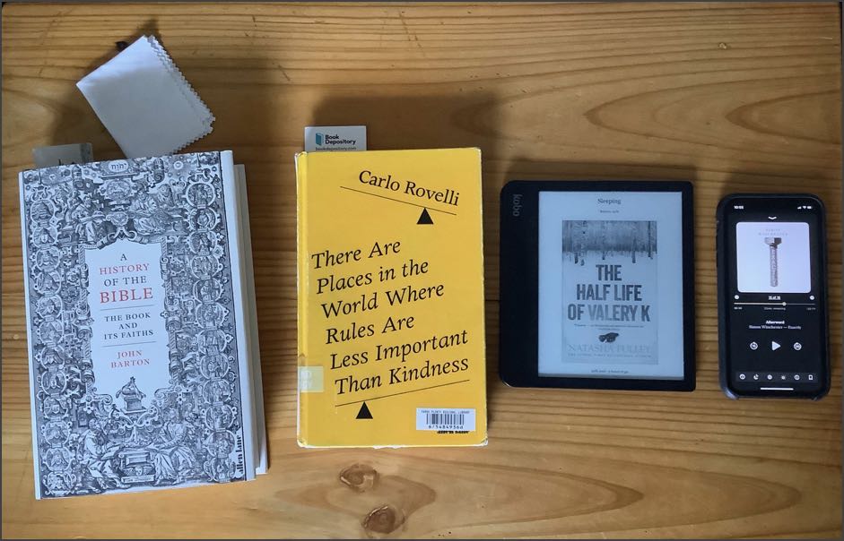 My ‘Reading Table’, showing the covers of four books. ‘A History of the Bible’ in hardcover, ‘There Are Places in the World Where Rules Are Less Important Than Kindness’ in a hardcover from the library, ‘The Half Life of Valery K’ on an ereader and ‘Exactly’ as an audiobook on a phone